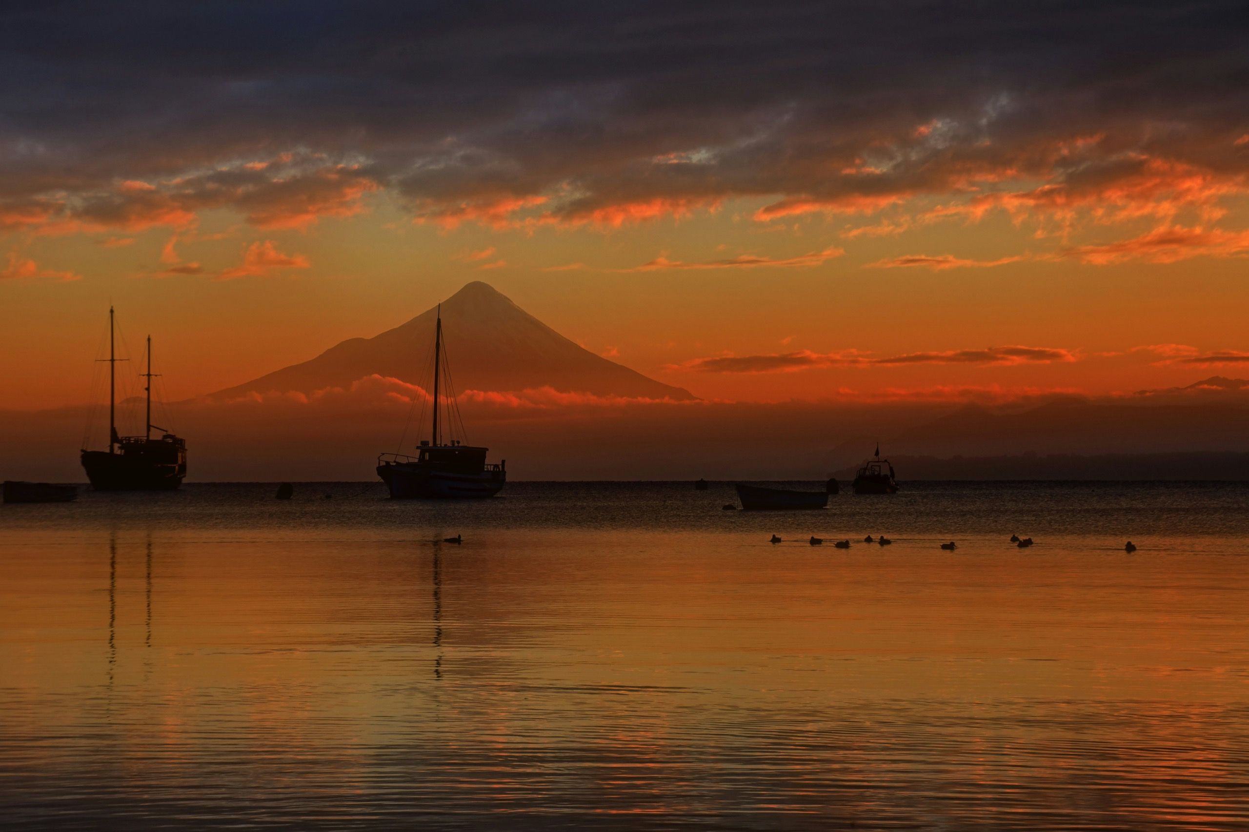El volcán Osorno recortado contra un cielo naranja al atardecer, con veleros fondeados y aves sobre el agua espejada del lago Llanquihue